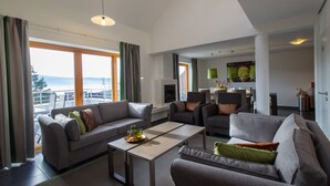 Un salon moderne avec un canapé, des fauteuils et une table basse. De grandes fenêtres offrent une vue sur l’extérieur.
