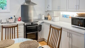 A modern kitchen with white cabinets, a stainless steel range hood, a built-in oven, a microwave, and a dining area with a table and chairs. - Coldwell Street Cottage (Wirksworth)
