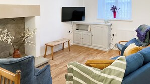 A living room with a blue sofa, a wooden bench, a television mounted on the wall, and a window with a plant. - Coldwell Street Cottage (Wirksworth)