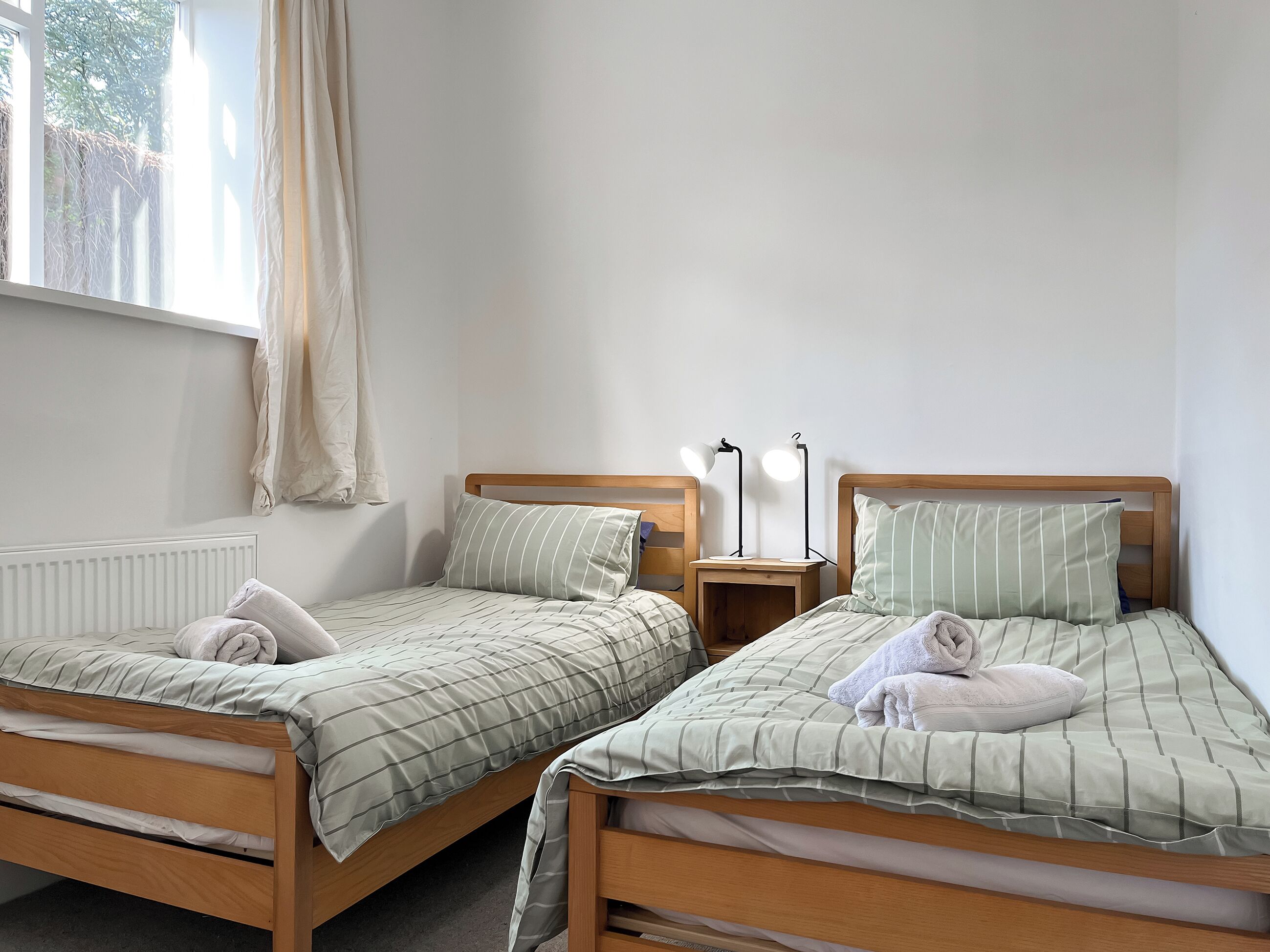 Two single beds with wooden frames and matching bedding, a bedside table with a lamp, and a window with a curtain.