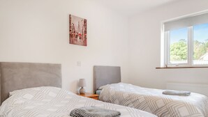 A bedroom with two beds, a nightstand with a lamp, a painting on the wall, and a window with a view of greenery. - An ideal destination for relaxation, comfort and immersion in nature’s beauty. (Old Buckenham)