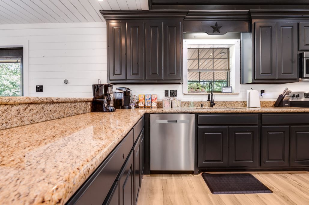 A modern kitchen with dark cabinetry, granite countertops, a stainless steel dishwasher, and a window with a view of greenery.