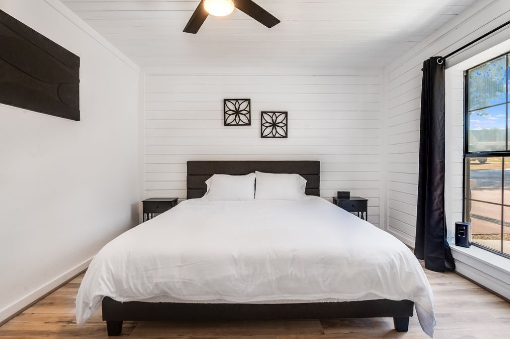 A bedroom with a large bed, a ceiling fan, two wall-mounted picture frames, and a window with a view of the outdoors.