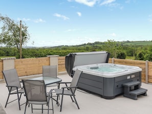 An outdoor hot tub with a table and chairs, surrounded by a wooden fence, overlooking a green landscape.
