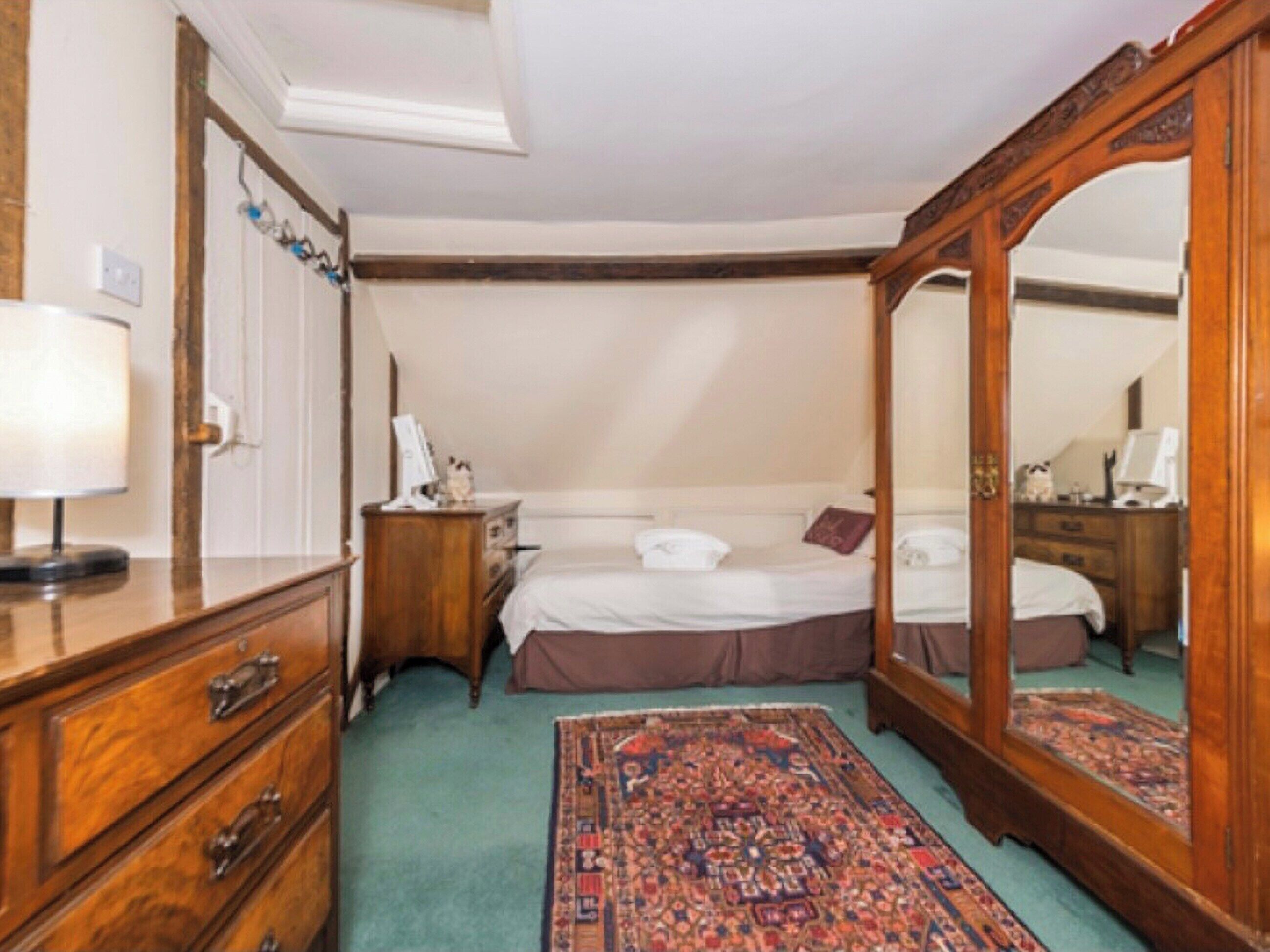 A traditional bedroom with a large mirror, a wooden dresser, a bedside table, and a patterned rug.