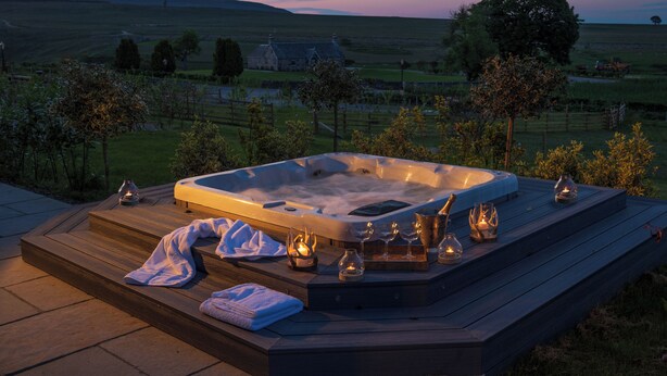 Un bain à remous avec des marches, entouré de bougies et de verres, installé sur une terrasse en bois offrant une vue panoramique sur la campagne environnante.