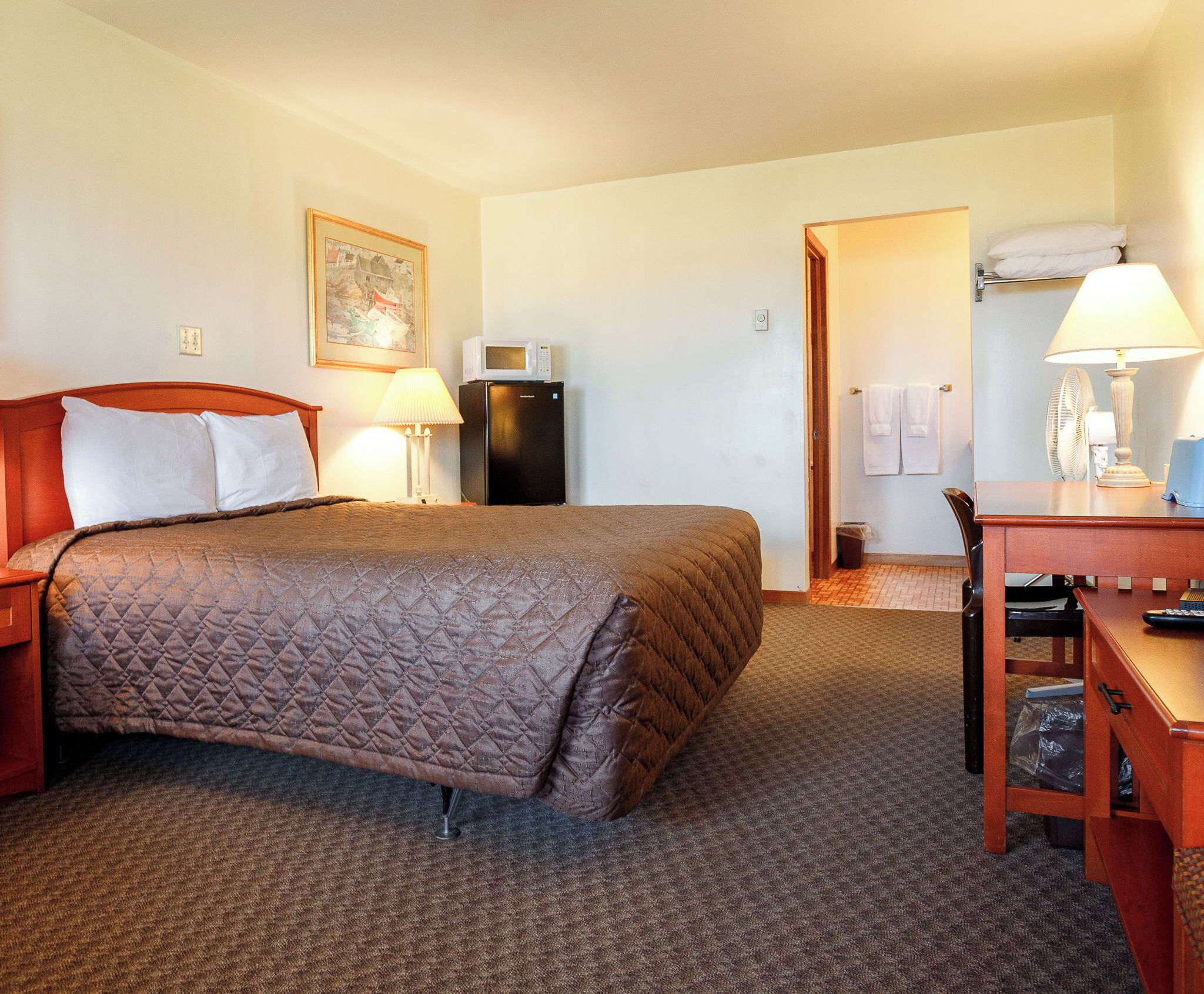 A hotel room with a bed, bedside lamp, refrigerator, and a desk with a lamp.