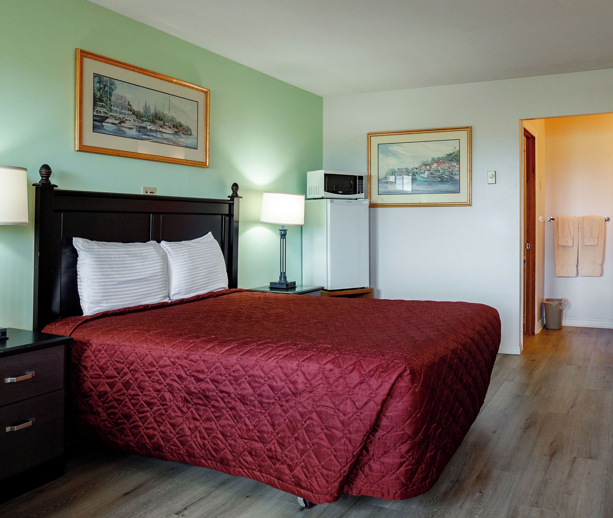 A neatly made bed with a red quilt, a nightstand with a lamp, a microwave, and two framed pictures on the wall.