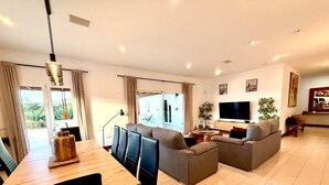 A modern living room with a dining area, grey sofas, a TV, and a large window with curtains. - Incredible 6 bed 5 bath detached villa in the exclusive area of Puerto Calero. (Puerto Calero)