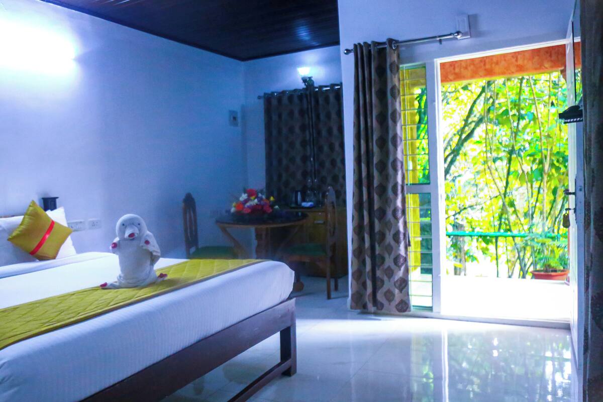 A hotel room with a bed, a small table, a chair, and a window with a view of greenery.