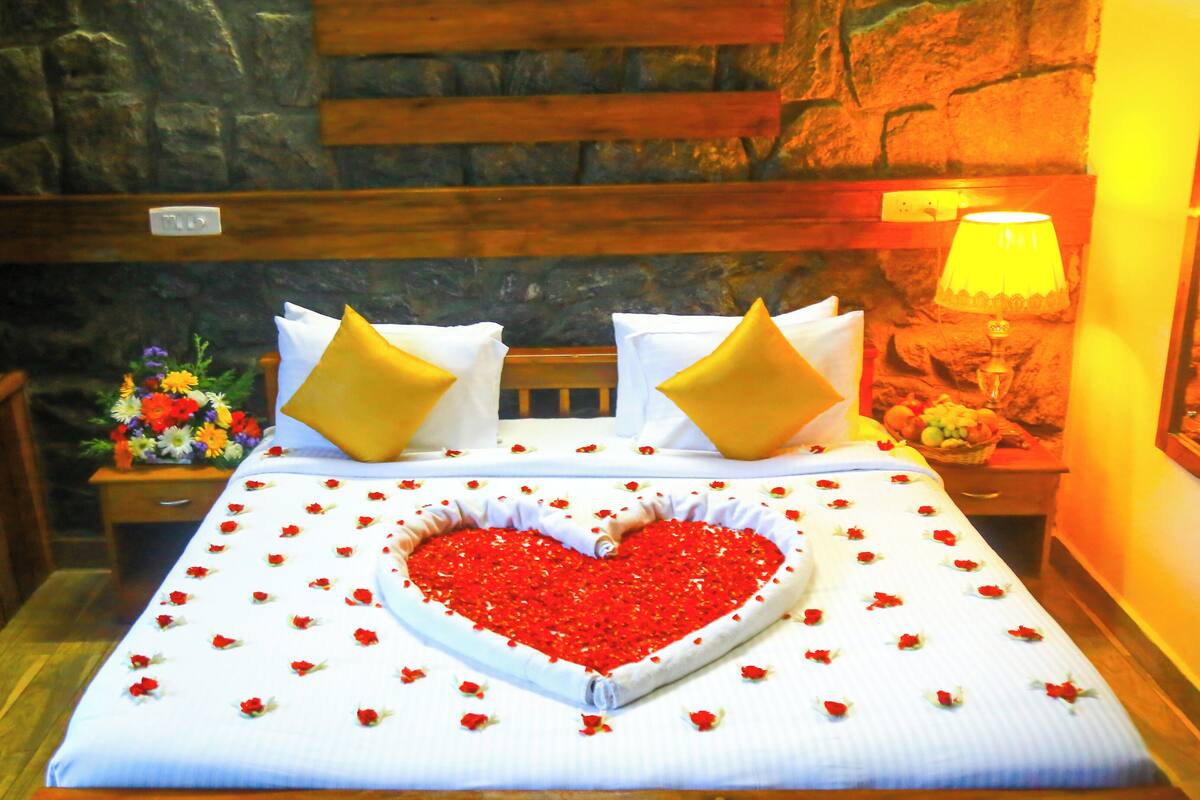 A bed with a heart-shaped arrangement of red flowers, two pillows, a nightstand with a vase of flowers, and a stone wall backdrop.
