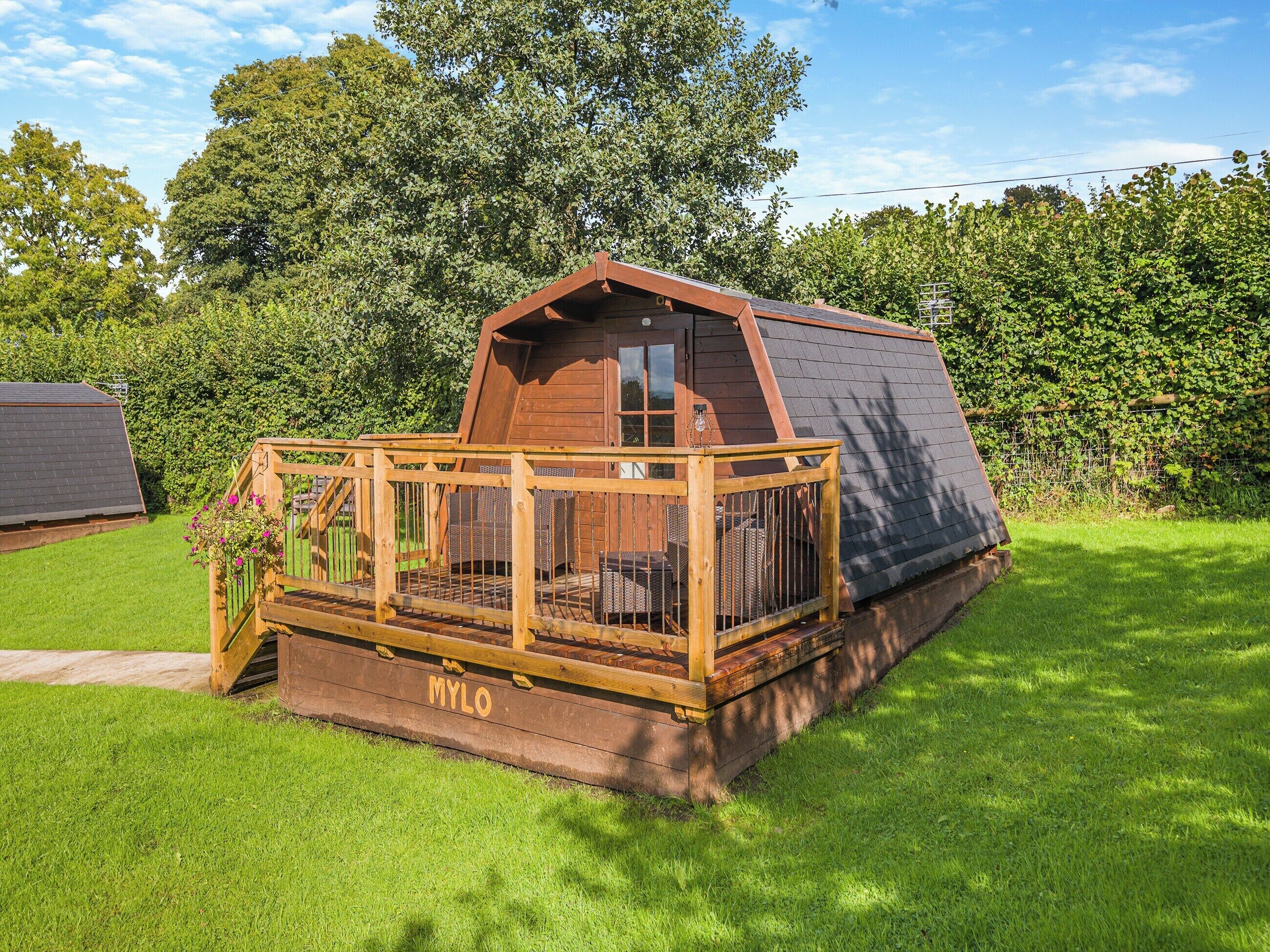 A wooden cabin with a sloped roof, a deck with wicker furniture, and a name 'MYLO' on the deck.