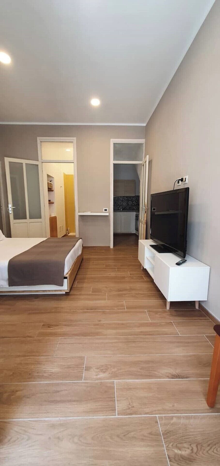 Une chambre d’hôtel avec un lit, un téléviseur posé sur un meuble blanc et un coin cuisine visible à travers une porte ouverte.