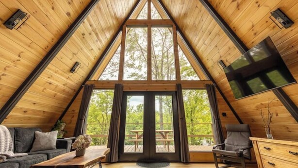 A cozy, triangular wooden interior with a large window, a sofa, a table, and a chair. - Thor’s Cabin luxury pet-friendly A-Frame w/ hot/cold tub & sauna 15 min to GSMNP (Cosby)