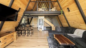 An attic-style room with a sloped ceiling, wooden flooring, a dining area with a table and chairs, a sofa, and a TV mounted on the wall. - Thor’s Cabin luxury pet-friendly A-Frame w/ hot/cold tub & sauna 15 min to GSMNP (Cosby)