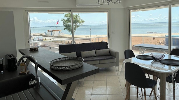 Una cuina moderna amb un gran finestral amb vista a una platja, zona de menjador amb taula i cadires, i un sofà al centre.