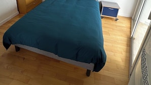 Un dormitori amb un llit gran, un taulell de nit amb una làmpada, un prestatge de fusta i un petit armari blau.