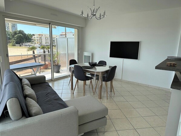 Un salon moderne avec un canapé, une table à manger et des chaises. Un téléviseur est fixé au mur et un balcon offre une vue sur les bâtiments et les arbres.