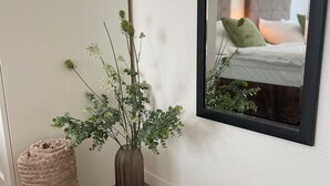 Canto com um vaso castanho com folhagem verde, uma almofada bege e um espelho refletindo uma cama com almofadas.