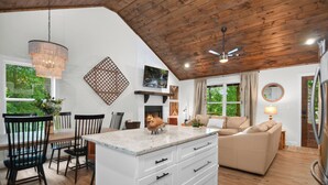 Amplia sala de estar con techo de madera, una isla blanca con cajones, un sofá color beige y un comedor con sillas y mesa.
