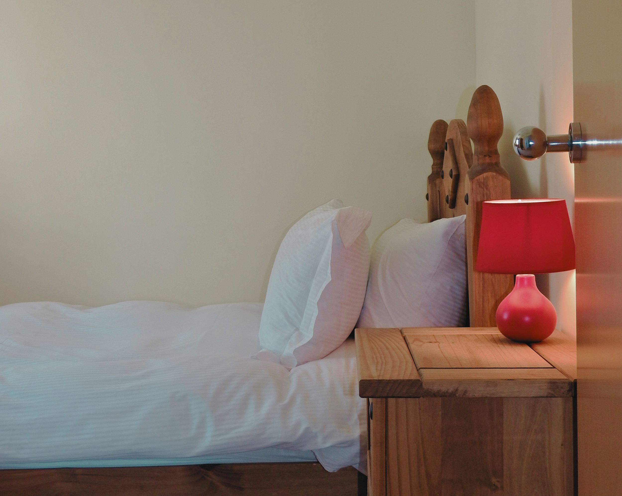 A bed with white linens, a wooden nightstand with a red lamp, and a wooden headboard.