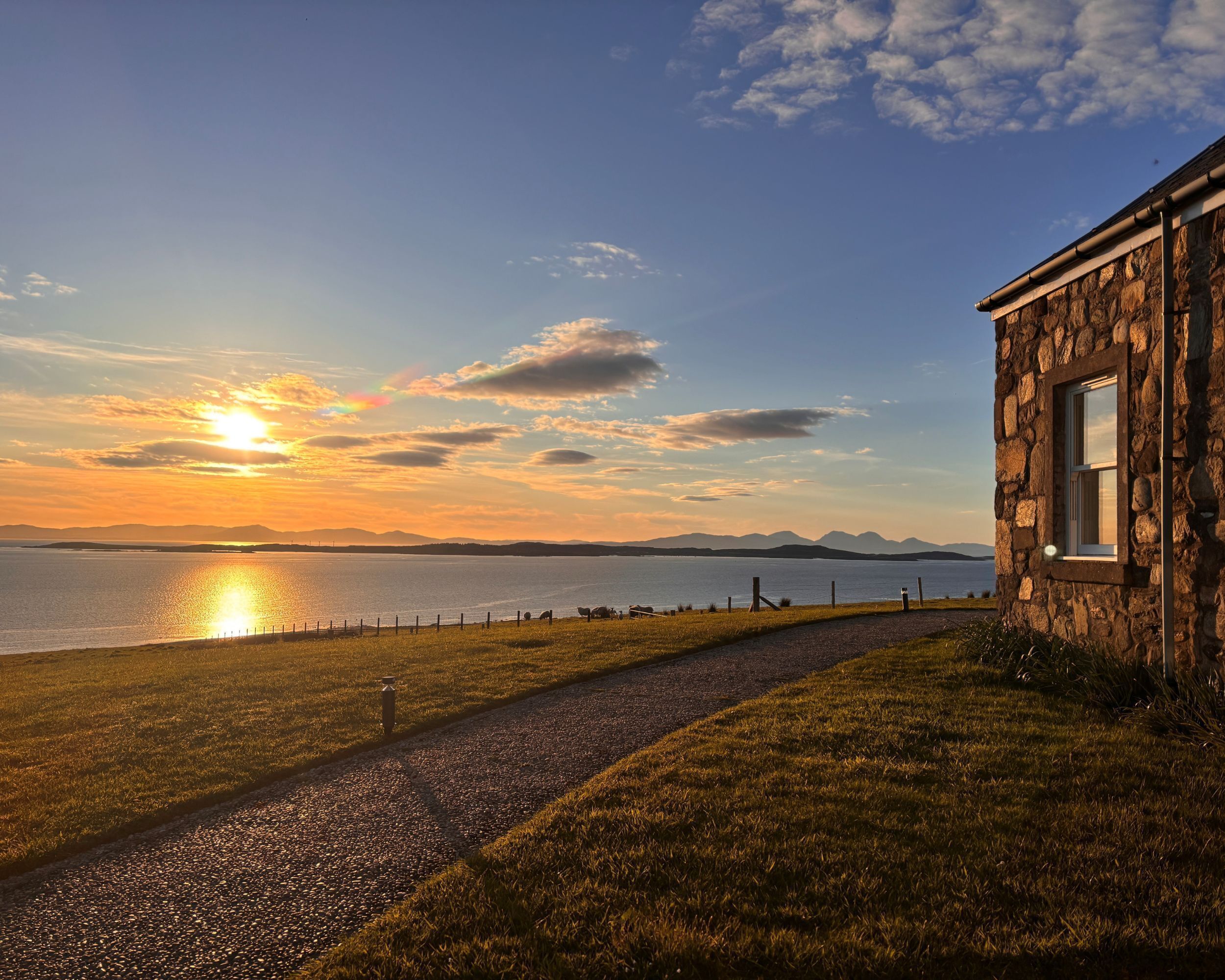 Ein Steinhaus mit einem Fenster, einem Kiesweg und Blick auf den Sonnenuntergang über dem Wasser.
