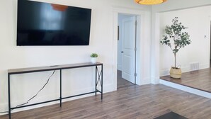 A modern living room with a large flat-screen TV mounted on the wall, a wooden console table, a potted plant, and a wicker pendant light.