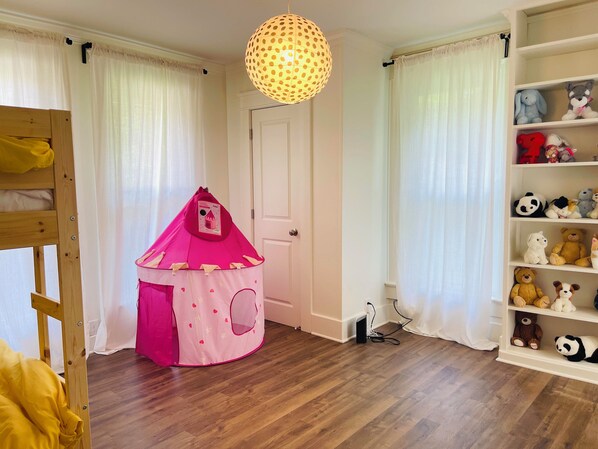 A child's play tent in a room with wooden flooring, a shelf with stuffed animals, and a hanging light fixture.