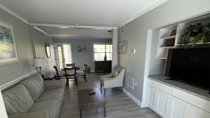 A living room with a sofa, a striped armchair, a coffee table, and a television. There is a dining area with a table and chairs, and a kitchen with cabinets and a countertop. - Lakeview Superior Two-Bedroom Suite (Marco Island)