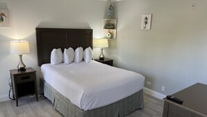 A neatly made bed with a headboard, two bedside tables with lamps, a small shelf with decorative items, and a remote control on a cabinet. - Lakeview Superior Two-Bedroom Suite (Marco Island)