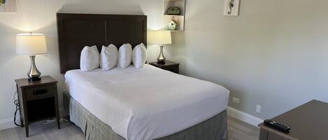 Una cama bien hecha con cabecera, dos mesitas de noche con lámparas, un estante con adornos y un control remoto sobre un mueble.