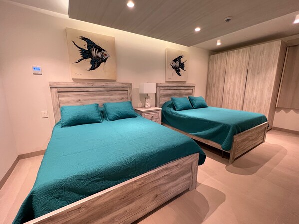 Une chambre avec deux lits, chacun agrémenté de linge de lit turquoise, de têtes de lit en bois et d’une table de chevet avec une lampe. Deux tableaux représentant des poissons sont accrochés au mur.