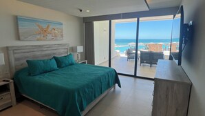 Une chambre dotée d’un grand lit, d’une table de chevet, d’une lampe, d’un tableau représentant une scène de plage, et offrant une vue sur l’océan et la piscine grâce à une porte-fenêtre coulissante.