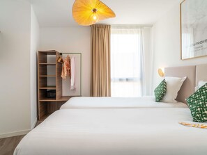 Une chambre d’hôtel comprenant un lit, une armoire, un porte-manteau, une fenêtre avec des rideaux et un luminaire fixé au mur.
