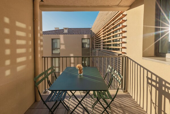 Een balkon met een tafel, stoelen, een bloempot en uitzicht op omliggende gebouwen.