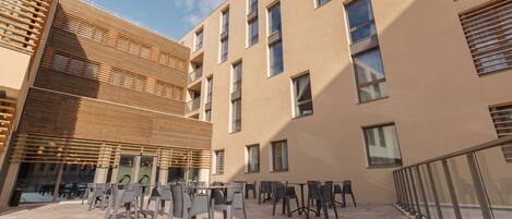 A modern building with multiple floors, outdoor seating area with tables and chairs, and a glass railing along the edge.
