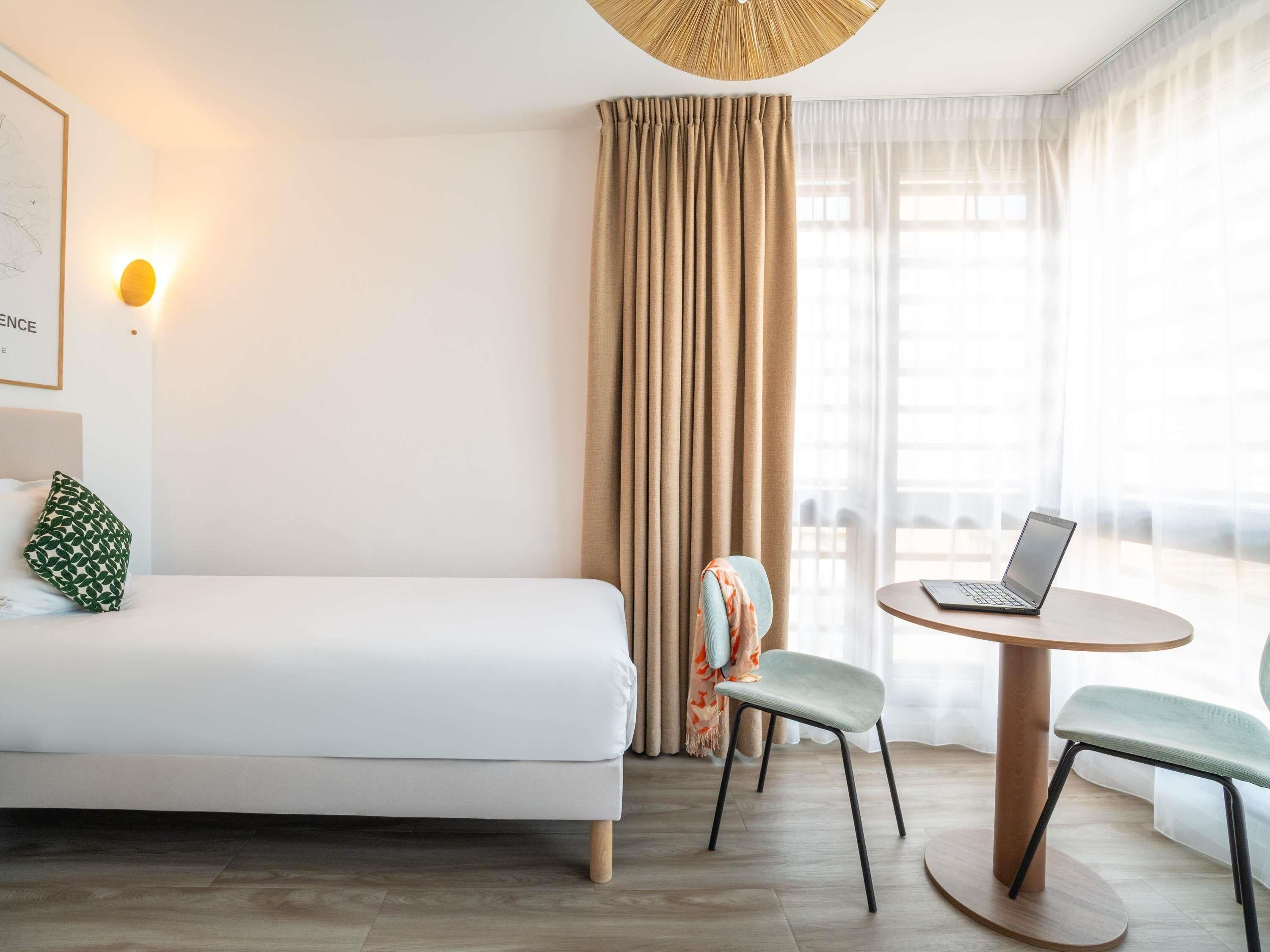 A hotel room with a bed, a desk with a laptop, a chair, and a large window with curtains.