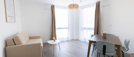 A modern living room with a sofa, a dining table, and chairs. There is a small round table with a vase of flowers. The room has large windows with curtains.