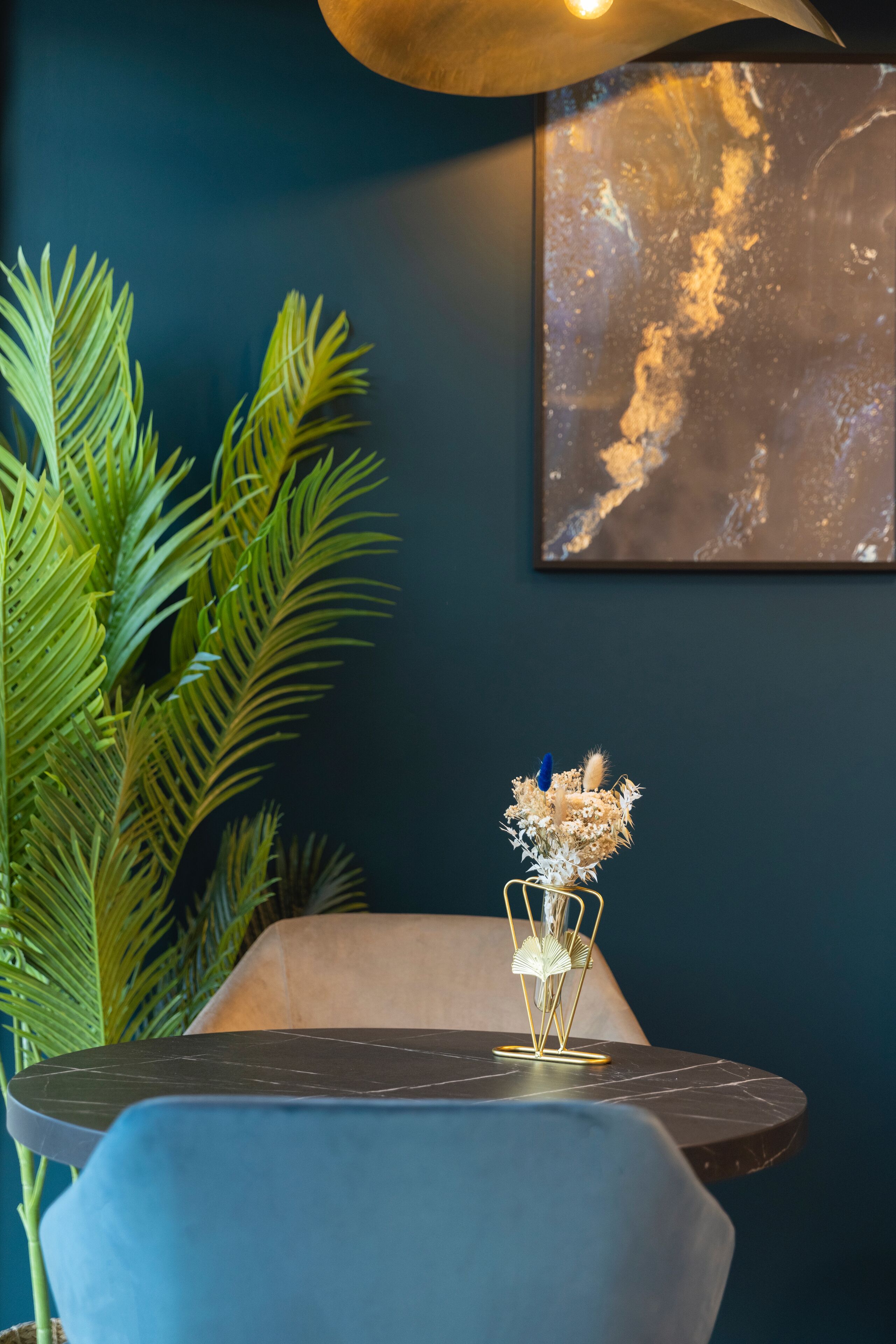 A room with a round table, a blue chair, a large potted plant, and a framed artwork on the wall.