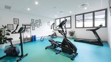 Ett gym med olika typer av träningsutrustning, inklusive löpband och en crosstrainer, på ett blått golv med vita väggar och inramade tavlor.
