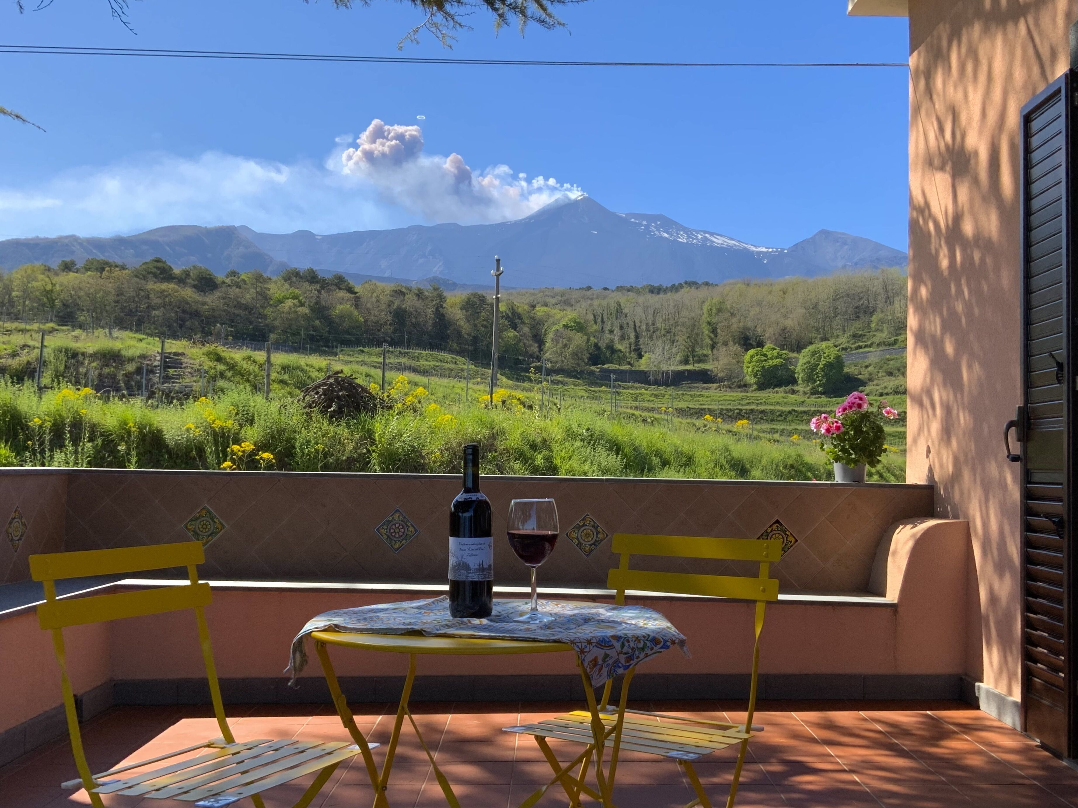 Uma varanda com uma mesa, cadeiras, uma garrafa de vinho e um copo. Ao fundo, vê-se uma montanha e um céu claro.