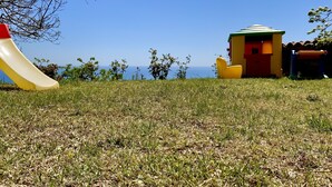 Une zone herbeuse avec un toboggan jaune, un petit bâtiment jaune et rouge, et une vue sur la mer.