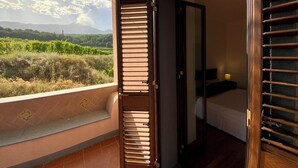 Un balcon donnant sur un vignoble et des montagnes, une porte en bois menant à une chambre avec un lit et une table de chevet, et un sol carrelé.