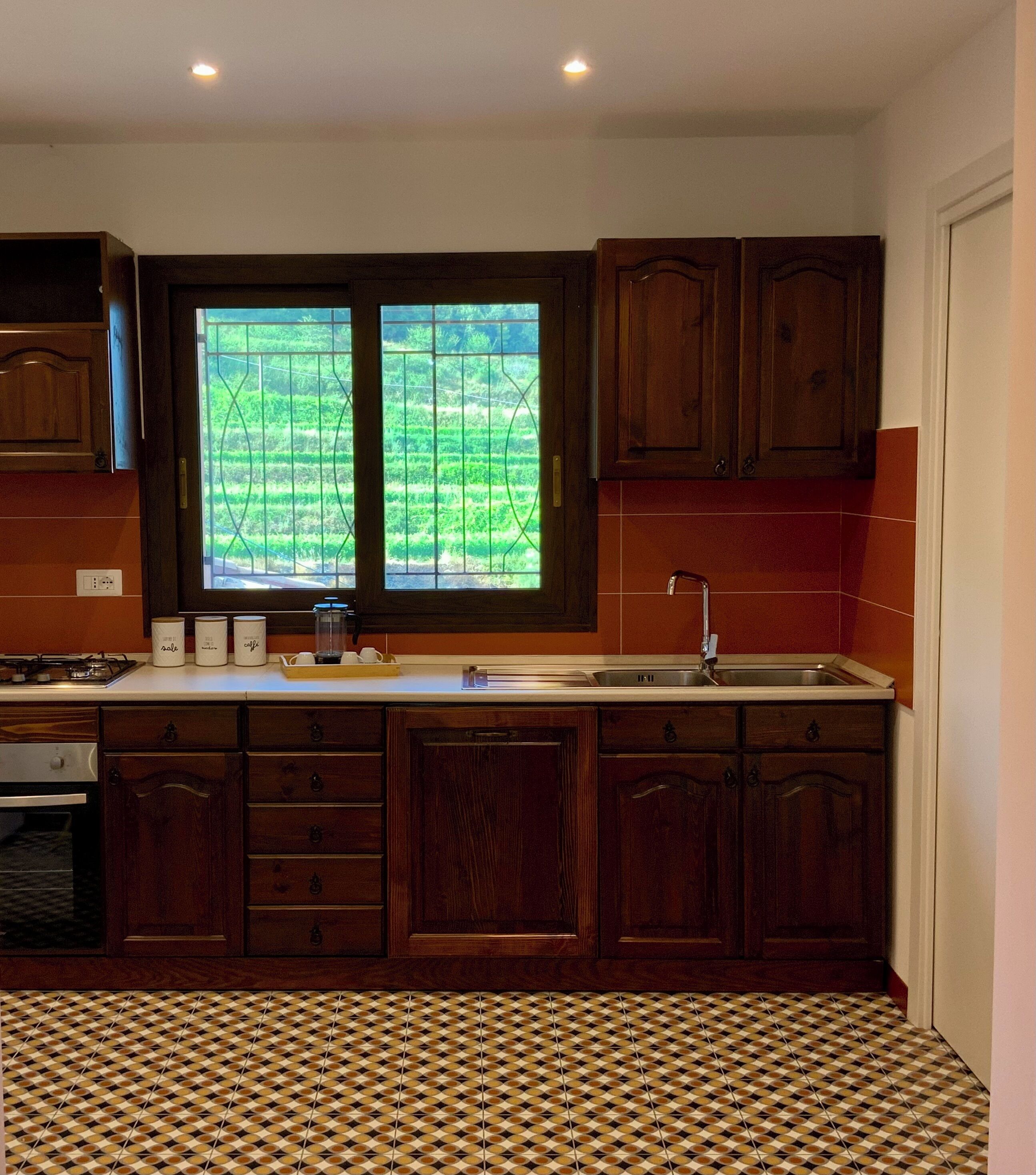Una cucina con mobili in legno, un'ampia finestra con vista su un campo verde, un lavandino e un pavimento decorato.