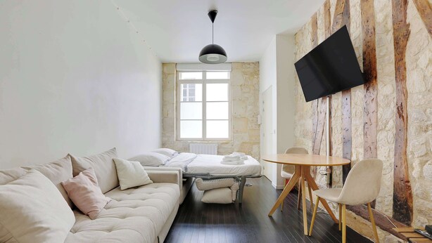 Quarto moderno com sofá, uma pequena mesa, TV fixada em uma parede com textura e janela que proporciona luz natural.