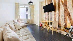 Uma sala de estar moderna com um sofá, uma mesa de jantar com cadeiras, uma televisão de tela plana fixada em uma parede texturizada e uma janela com vista para o exterior.