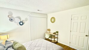 Quarto com cama, mesa de cabeceira com abajur, relógio de parede e porta.