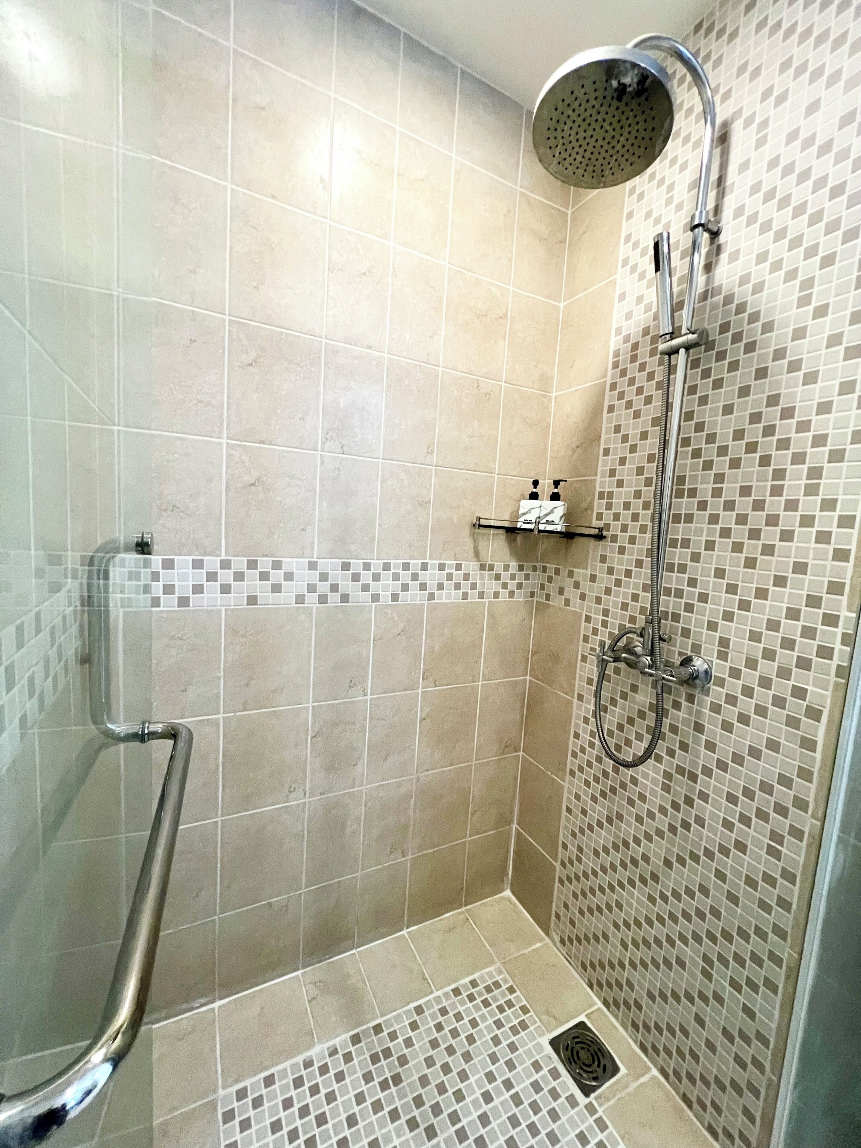 Zona de ducha con barra de apoyo, cabezal de ducha manual y pared de azulejos con diseño de mosaico.