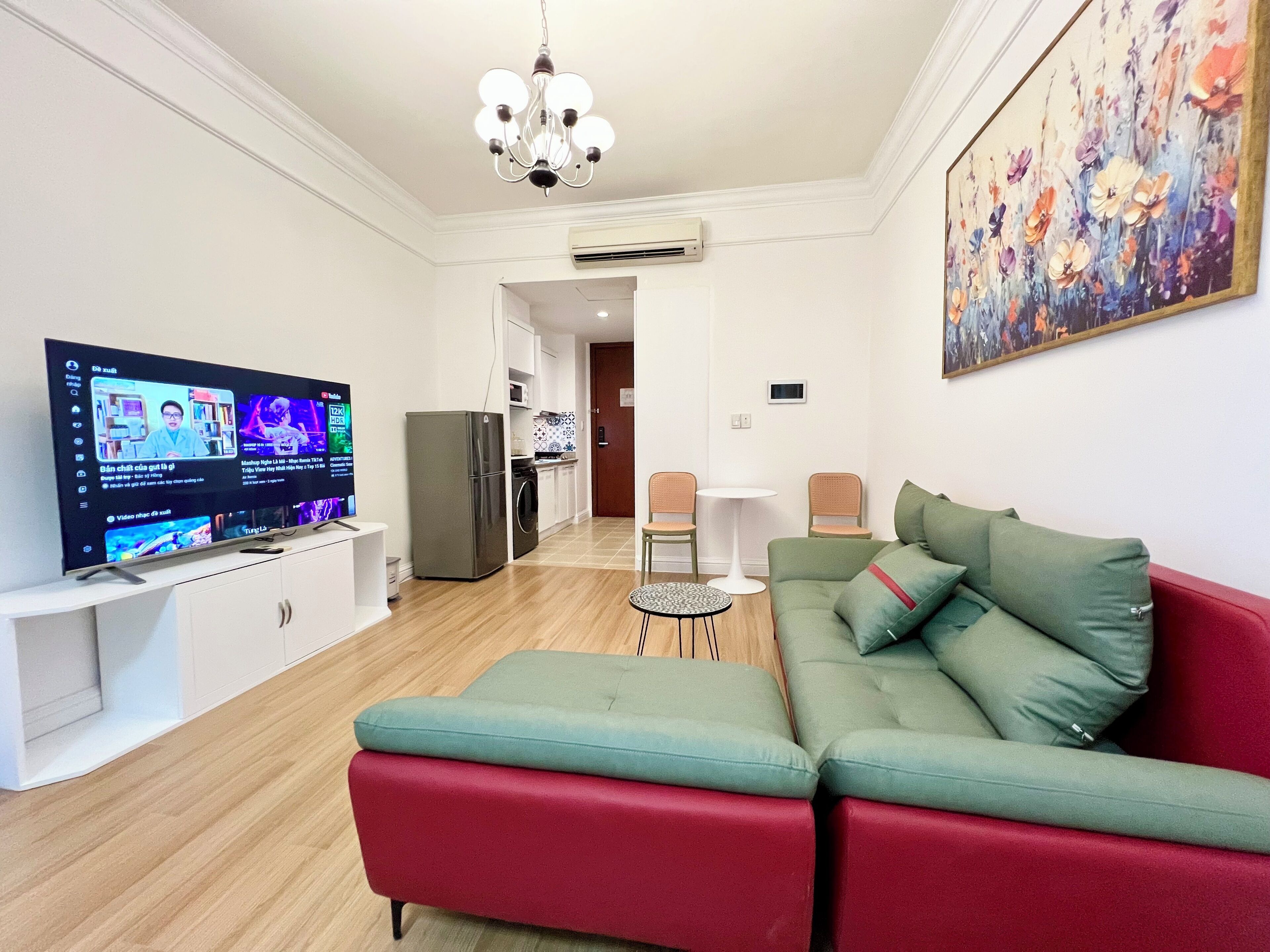 A modern living room with a flat-screen TV, a green sofa, a red sofa, a dining table, and a chandelier.