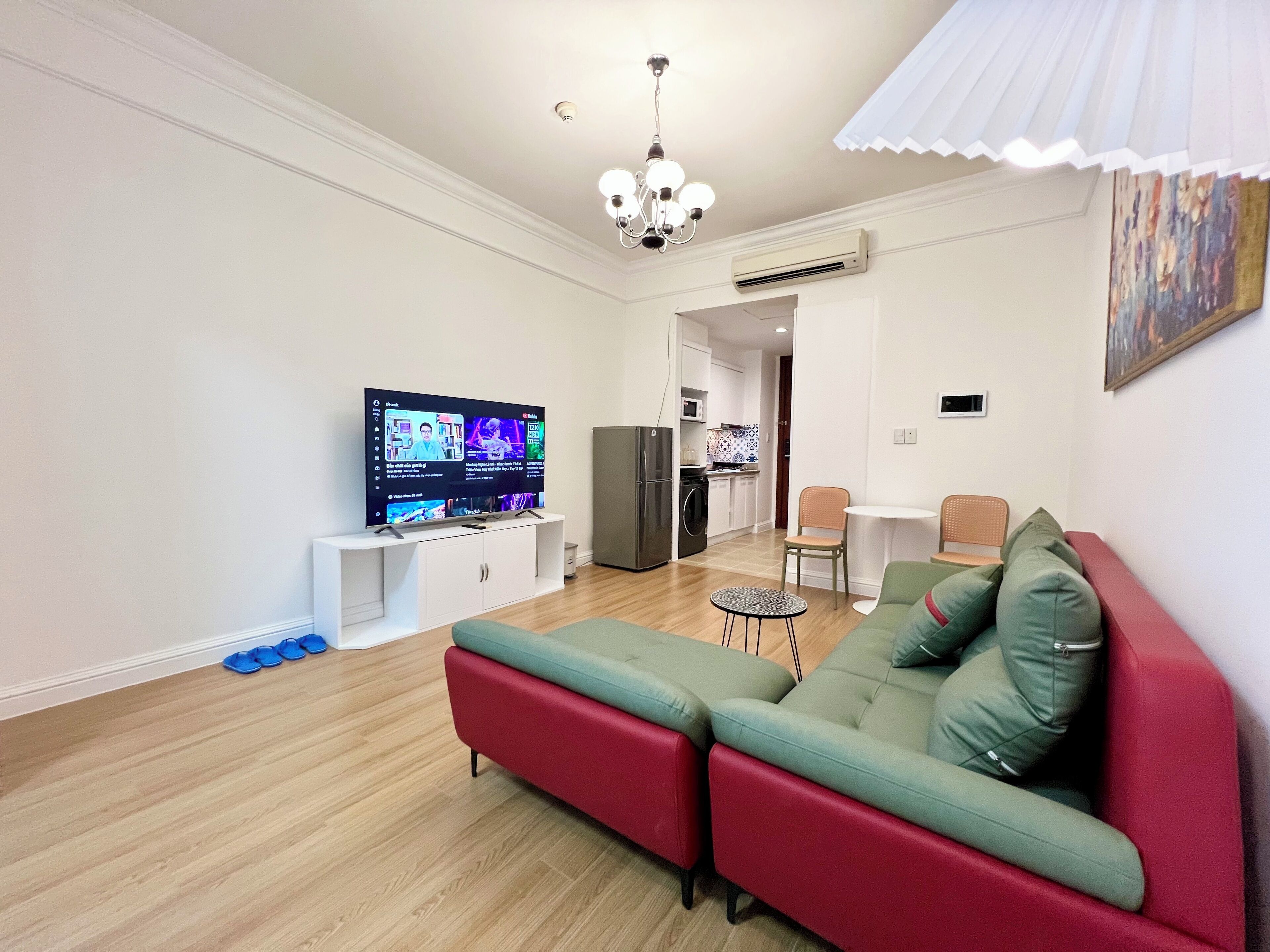 A modern living room with a flat-screen TV, a sofa, a small round table, and a kitchen in the background.